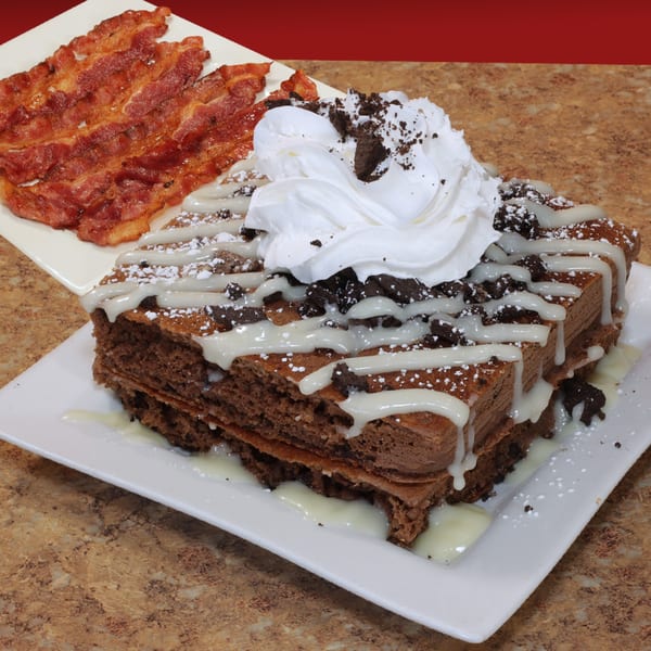 Chocolate chip pancakes with whipped cream, cookie crumbles, and bacon on a white plate.