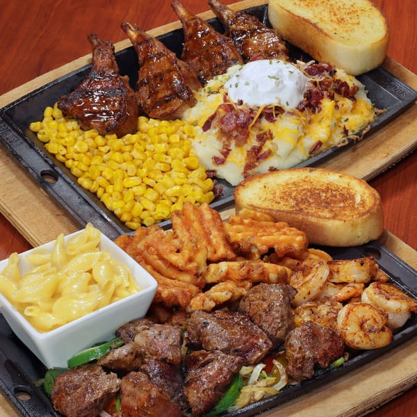 Two sizzling platters of grilled lamb chops, steak, shrimp, corn, mashed potatoes, waffle fries, and mac and cheese.