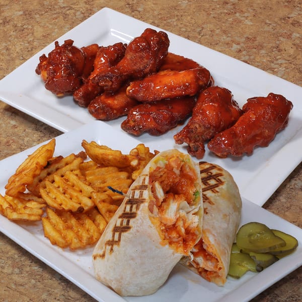 Plates of buffalo wings, a grilled chicken wrap with waffle fries, and pickles on a counter.